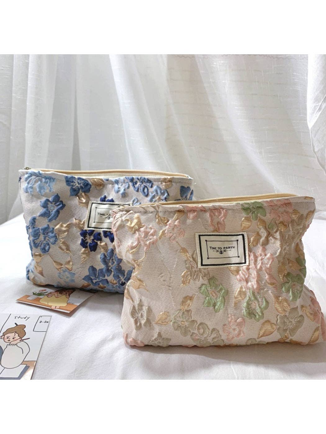 Two floral embroidered cosmetic pouches with gold-tone zippers, displayed on a white fabric surface in front of sheer white curtains. One pouch is light blue with blue floral embroidery, and the other is cream with pink and green floral embroidery.