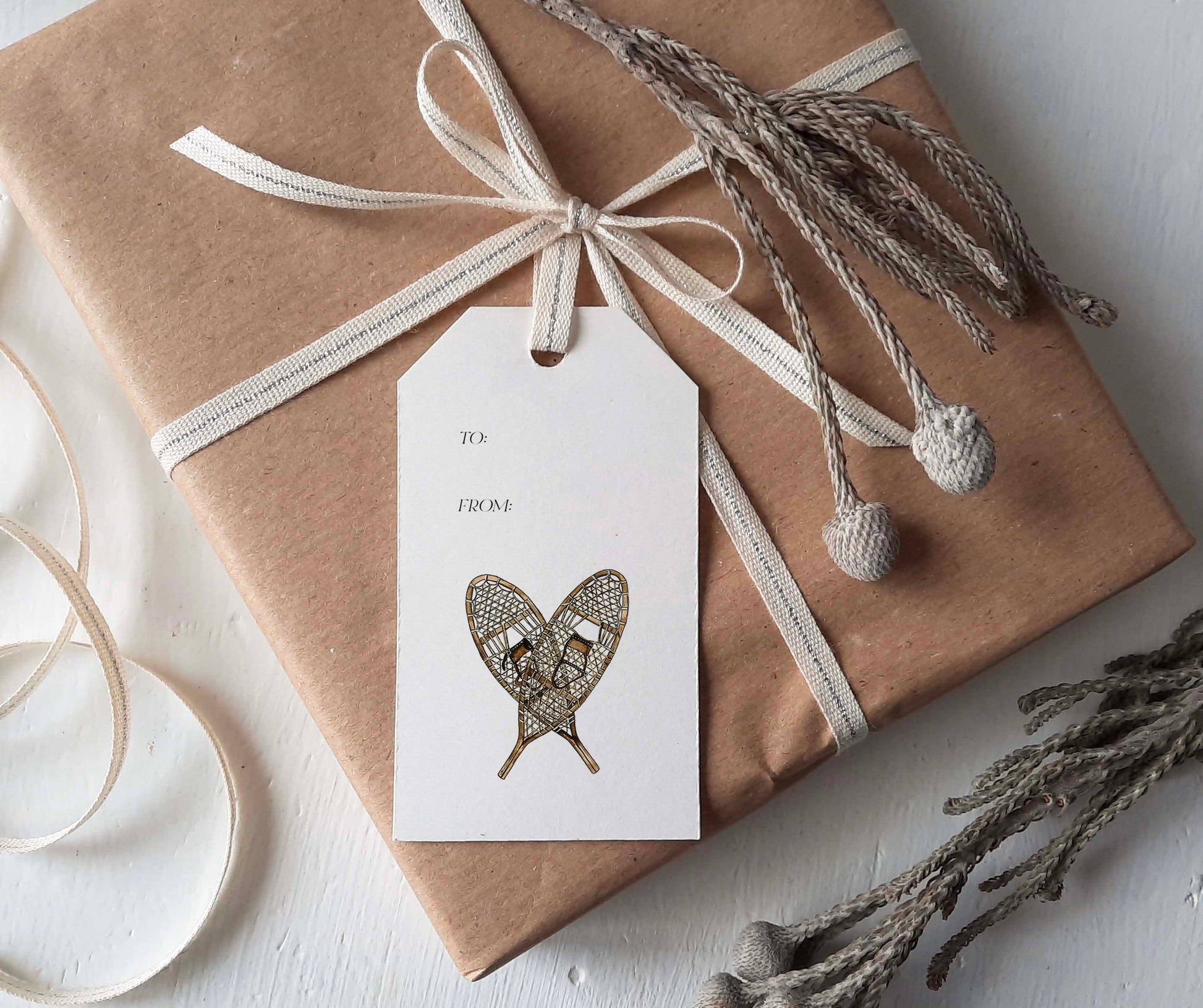 A wrapped gift box in brown kraft paper with a cream ribbon and bow, a white gift tag featuring a heart-shaped snowflake design, and dried decorative branches on a light surface.