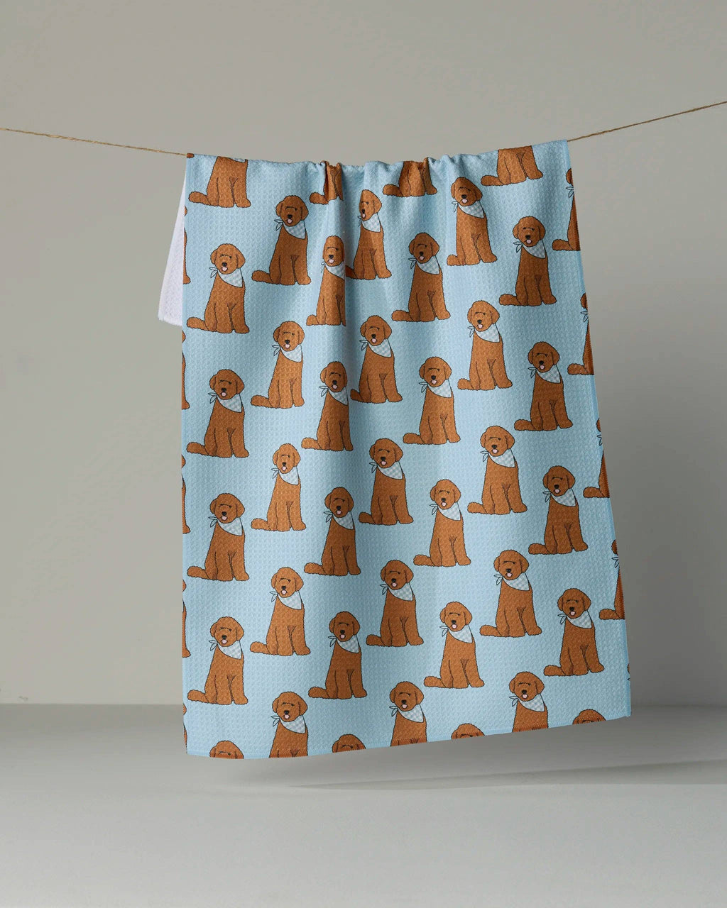 A light blue kitchen towel with an all-over repeating print of brown and white cartoon dogs, hanging from a string against a neutral gray background.