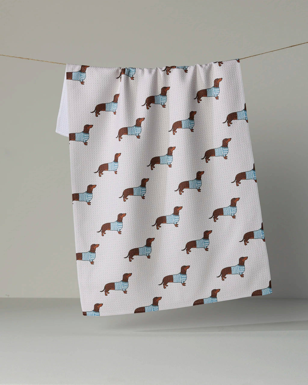 A white kitchen towel with a repeating pattern of dachshund dogs wearing blue sweaters, hanging from a string against a neutral gray background.