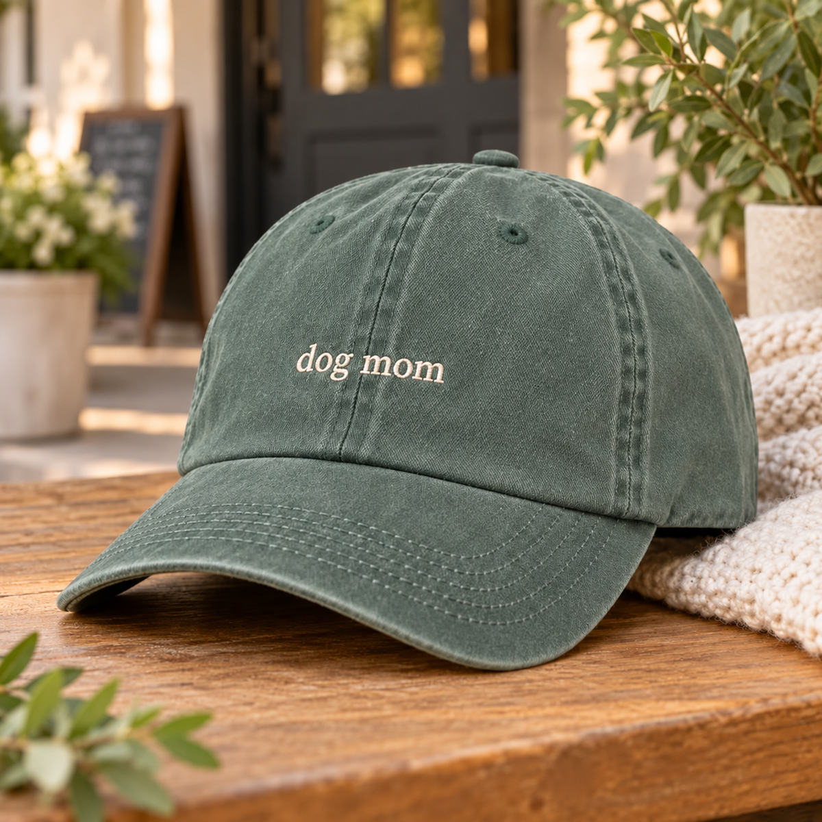 A dark green washed cotton baseball cap with 'dog mom' embroidered in white on the front, resting on a wooden table with a cozy knit blanket and blurred outdoor background.