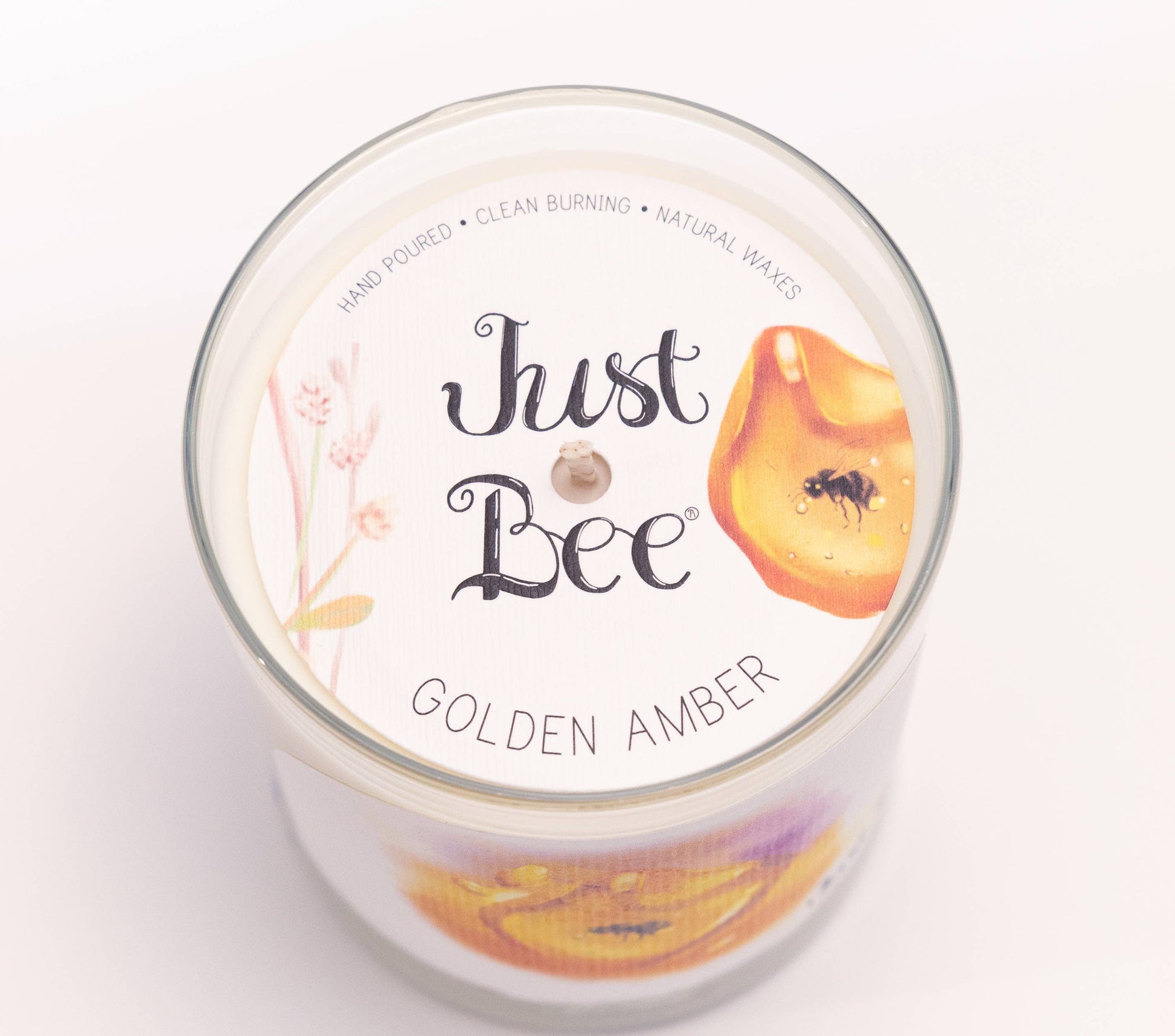A top-down view of a Just Bee Golden Amber scented candle in a clear glass jar, showing the white wax and printed label details.