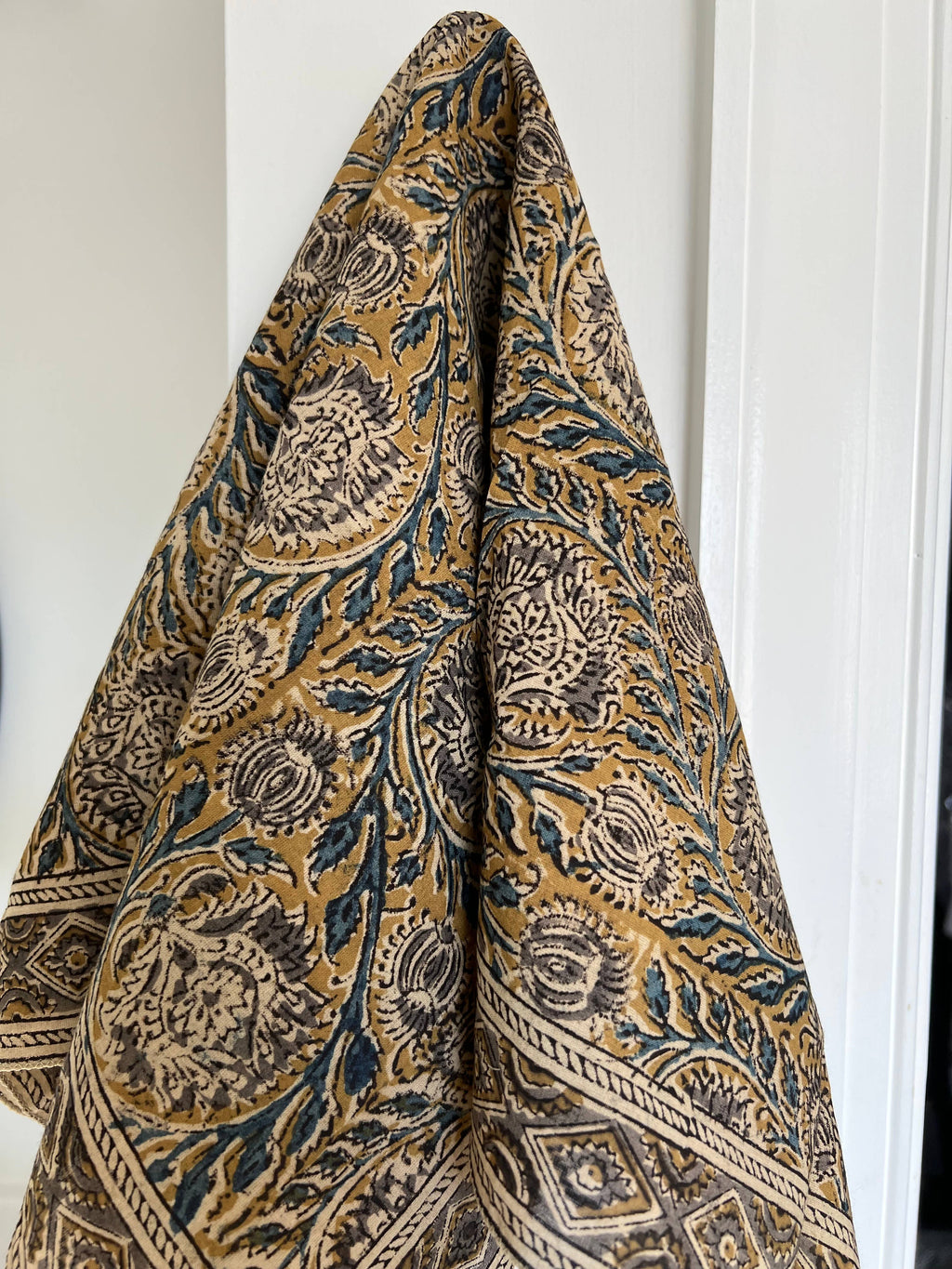 A folded patterned textile, likely a throw blanket or scarf, featuring a traditional block print design in mustard yellow, dark blue, and beige, hanging against a white paneled door.