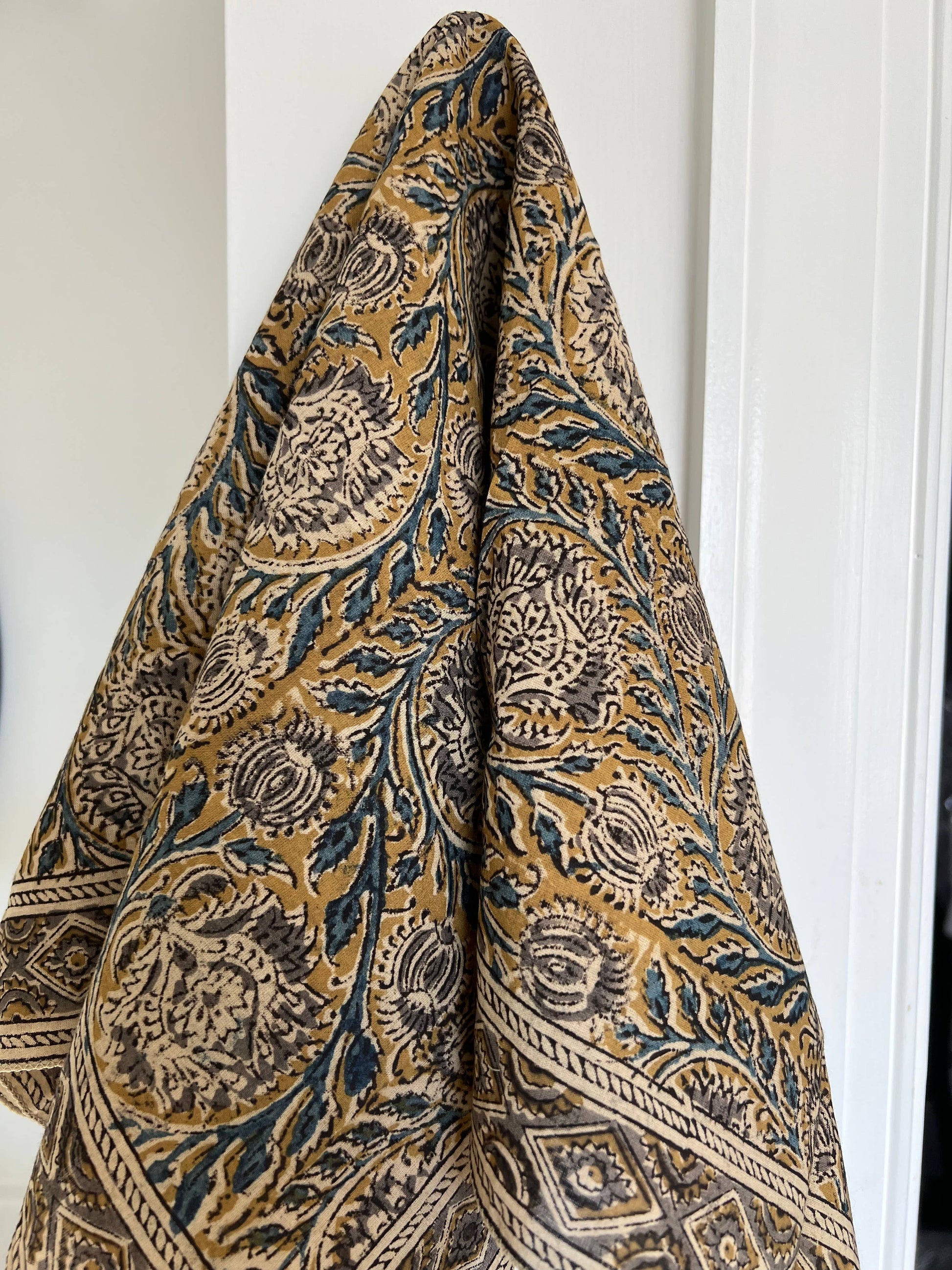 A folded patterned textile, likely a throw blanket or scarf, featuring a traditional block print design in mustard yellow, dark blue, and beige, hanging against a white paneled door.