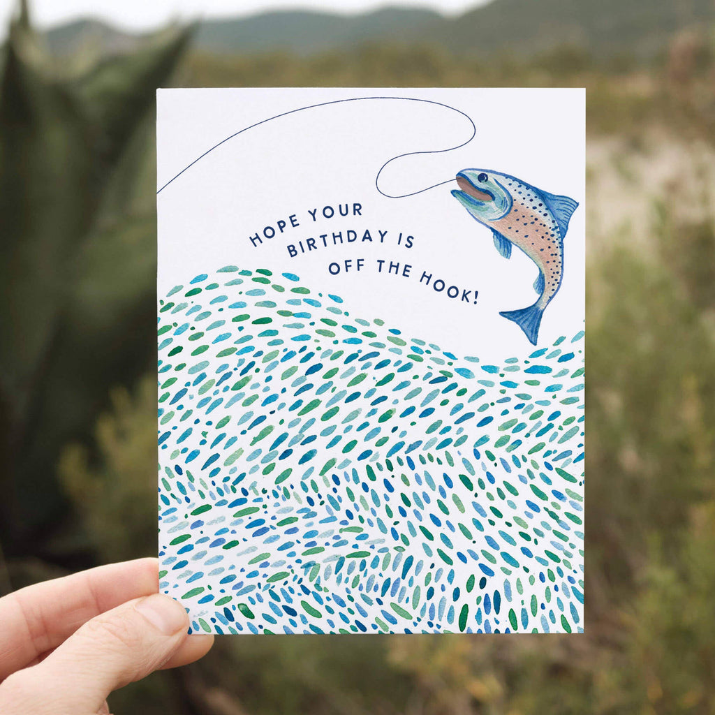 Hand holding a birthday card with a fish illustration and text against a natural background