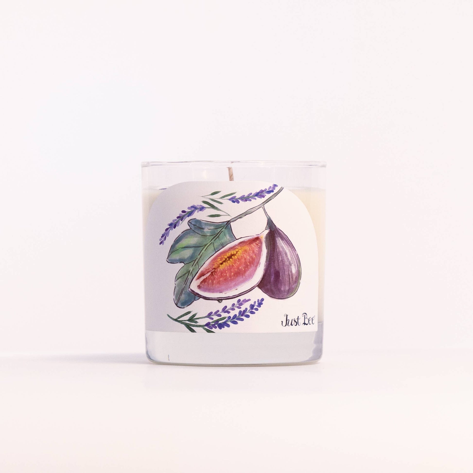 A clear glass jar candle with white wax and a printed label featuring a watercolor illustration of figs and lavender sprigs, photographed on a clean white background.