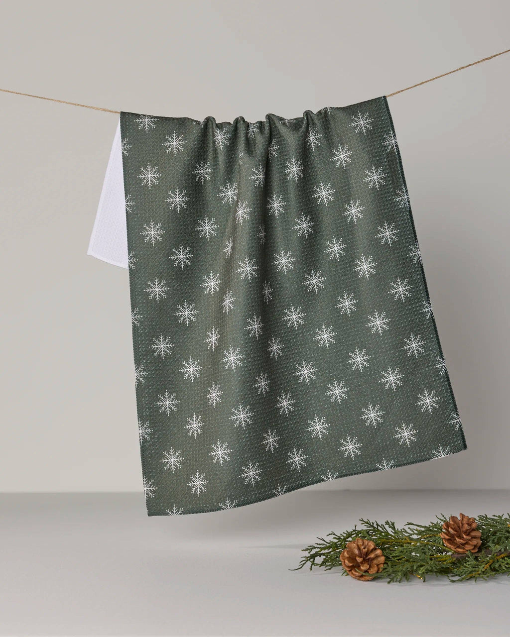 A dark green kitchen towel with a white snowflake pattern, hanging from a string in a studio setting with pine branches and pinecones at the bottom right.