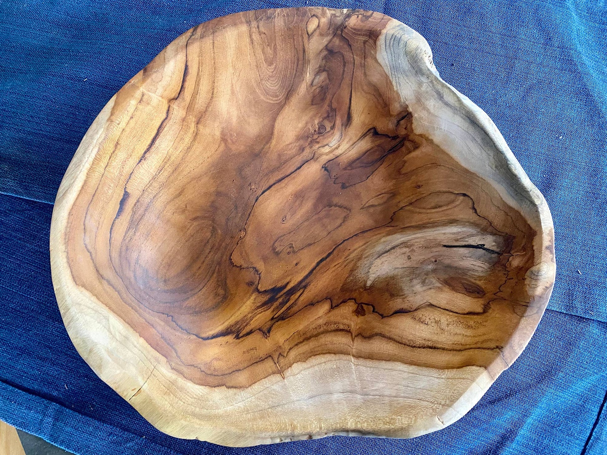 A rustic handcrafted wooden bowl with a live edge, showcasing natural wavy grain patterns in shades of brown and beige, set against a blue fabric background.