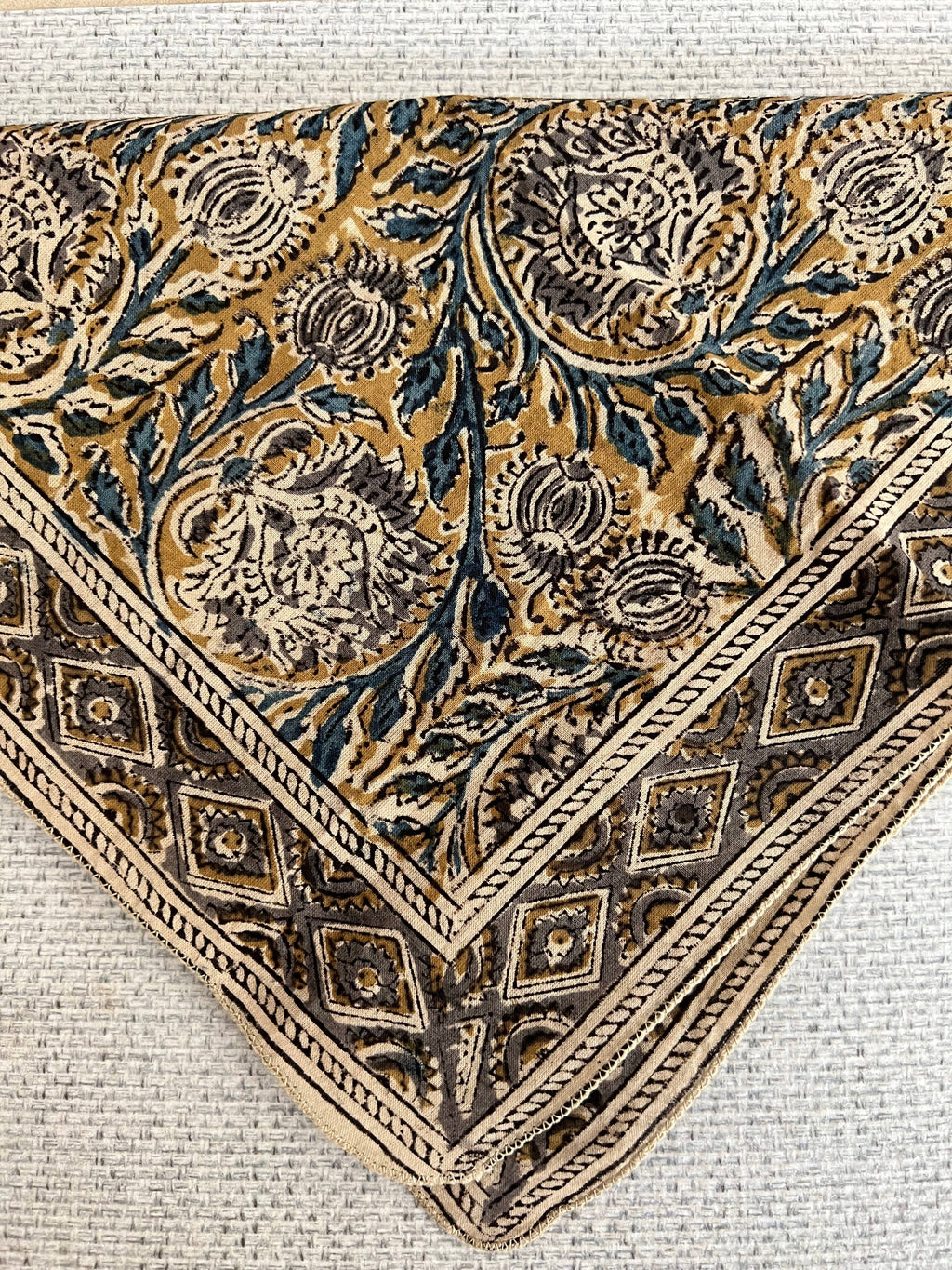 A close-up of a square bandana featuring an intricate floral and botanical pattern in gold, brown, and dark blue, finished with a detailed geometric border.