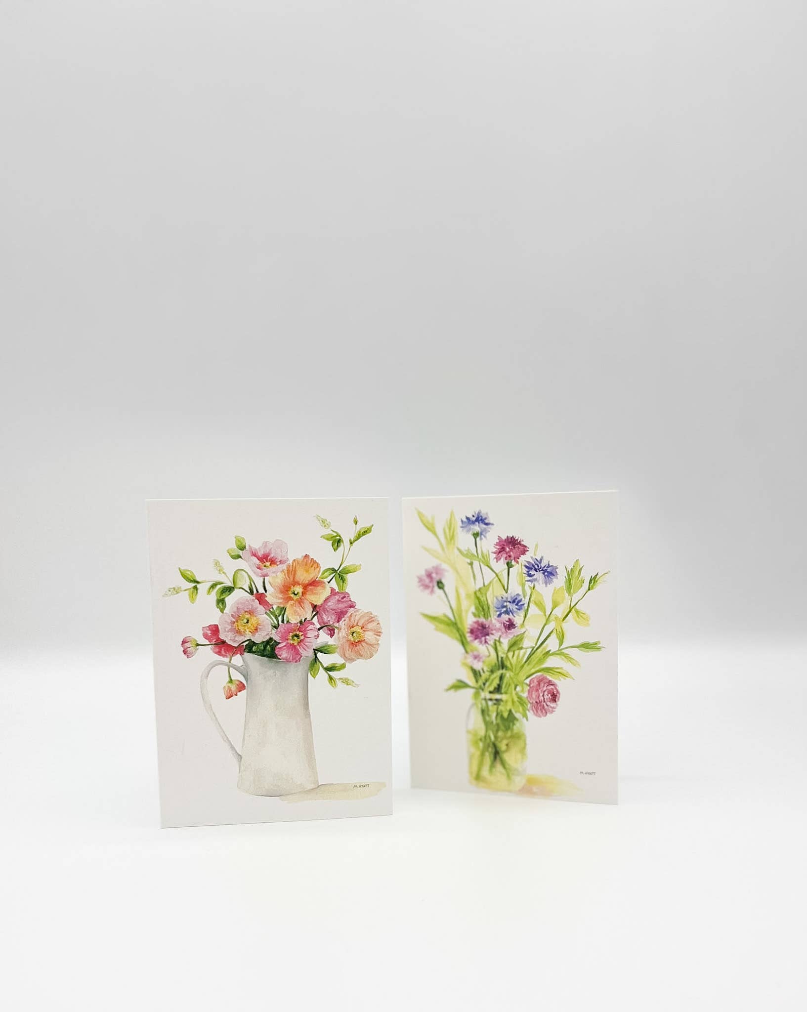 Two small greeting cards standing upright on a white surface against a light gray background. Each card features a watercolor-style floral bouquet in a vase: the left card shows pink, peach, and yellow flowers in a white pitcher, and the right card shows blue, pink, and purple flowers in a clear glass vase.