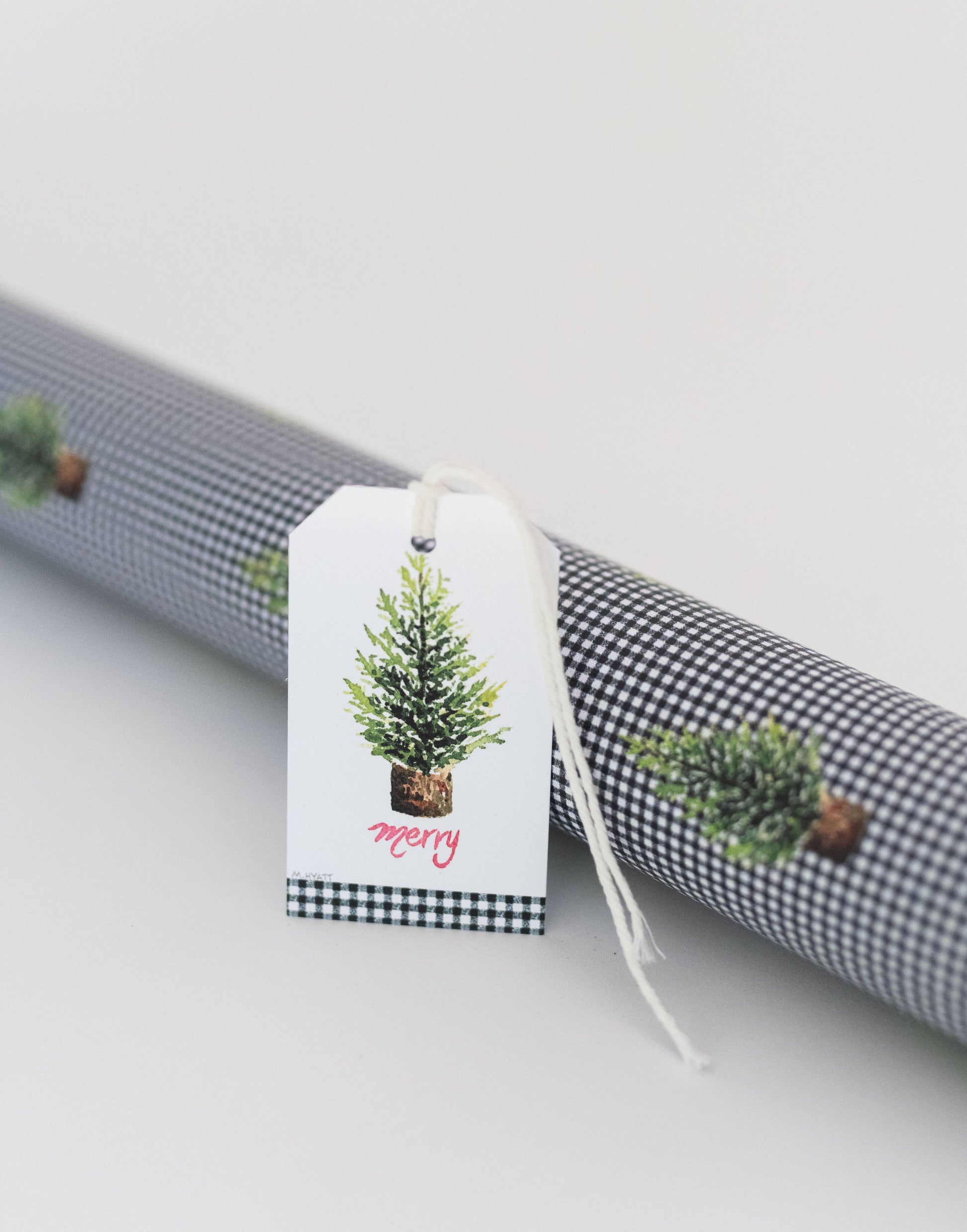 A white gift tag with a watercolor Christmas tree and the word 'merry' in red script, hanging from a roll of black-and-white gingham wrapping paper with small pine tree prints.