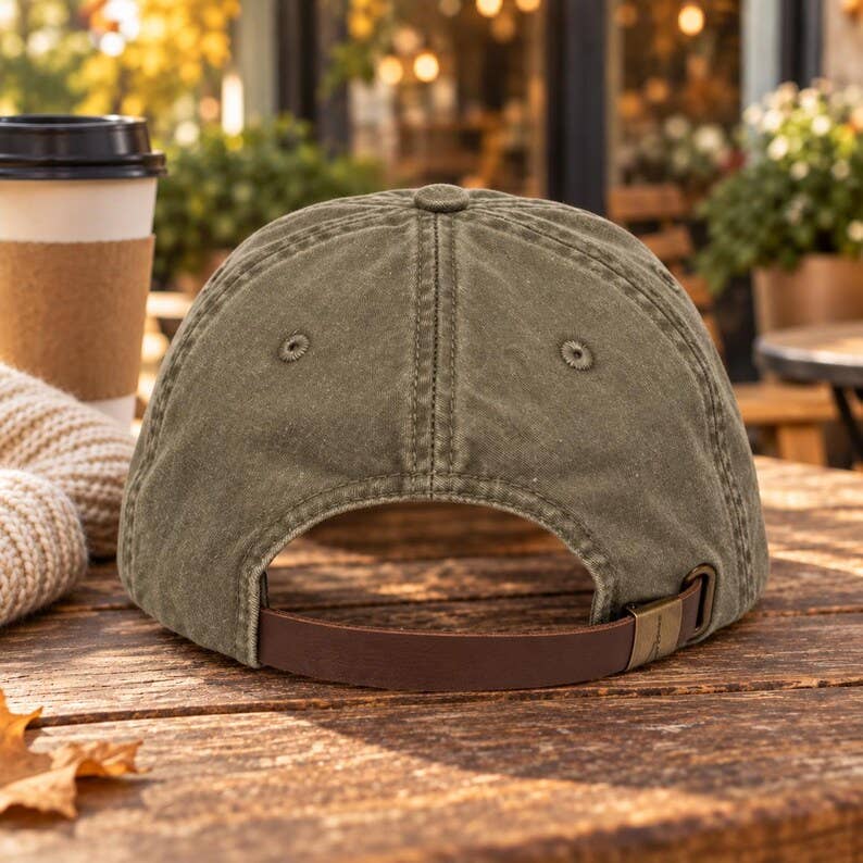 An olive green canvas baseball cap with a brown adjustable strap, shown from the back on a rustic wooden table next to a coffee cup in an outdoor cafe setting.