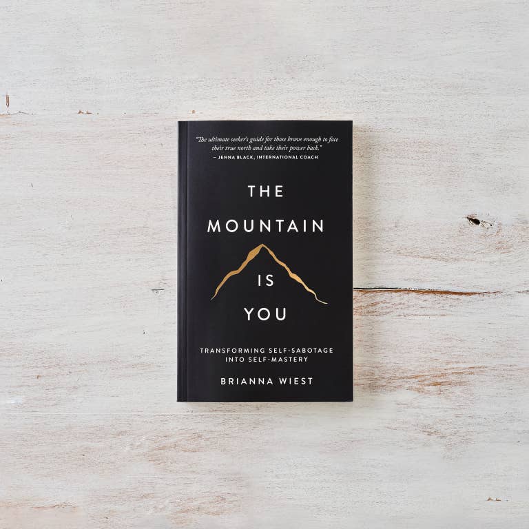 The book 'The Mountain Is You' by Brianna Wiest, featuring a black cover with white text and a gold mountain graphic, displayed on a light, rustic white wood background.
