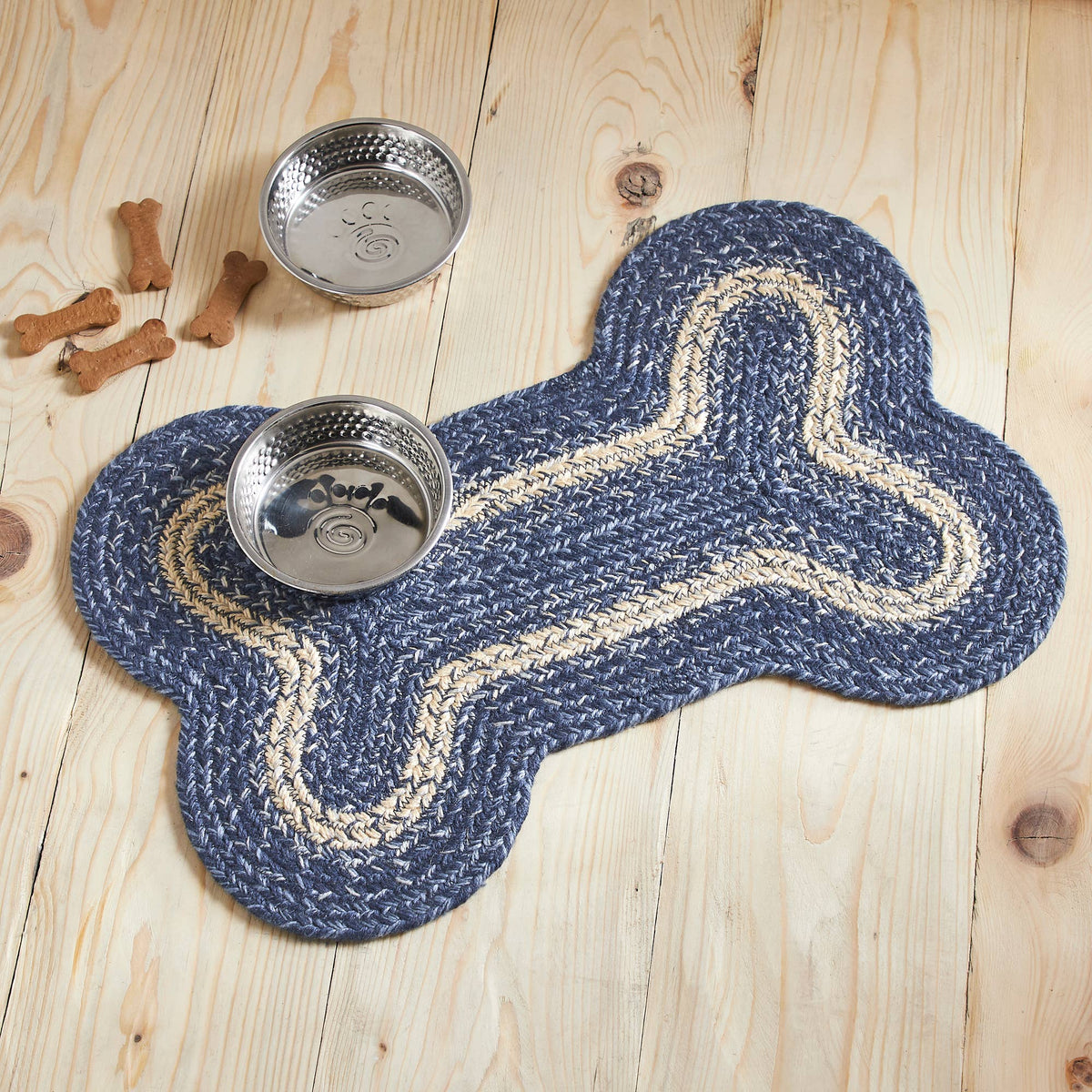A bone-shaped braided rug in navy blue and cream is placed on a light wood floor, with two stainless steel bowls and a few dog treats nearby.