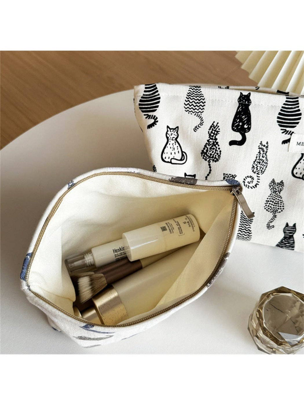 A white canvas cosmetic pouch with a black cat pattern, shown open with makeup items inside, placed on a white surface with a wooden background.