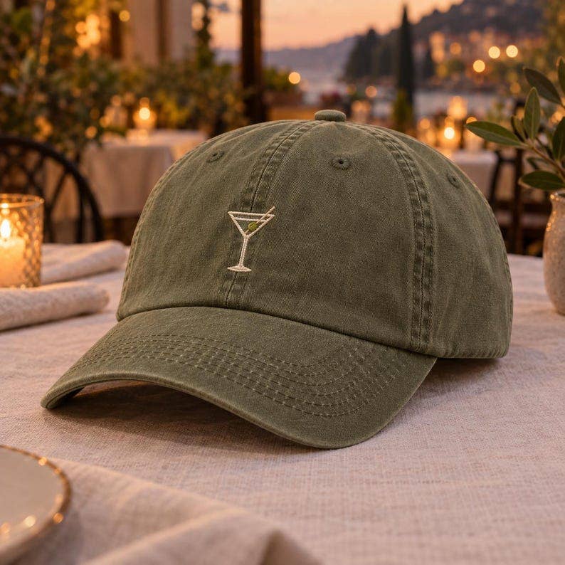 An olive green baseball cap with an embroidered martini glass logo on the front, resting on a table in a warm, softly lit outdoor dining setting.