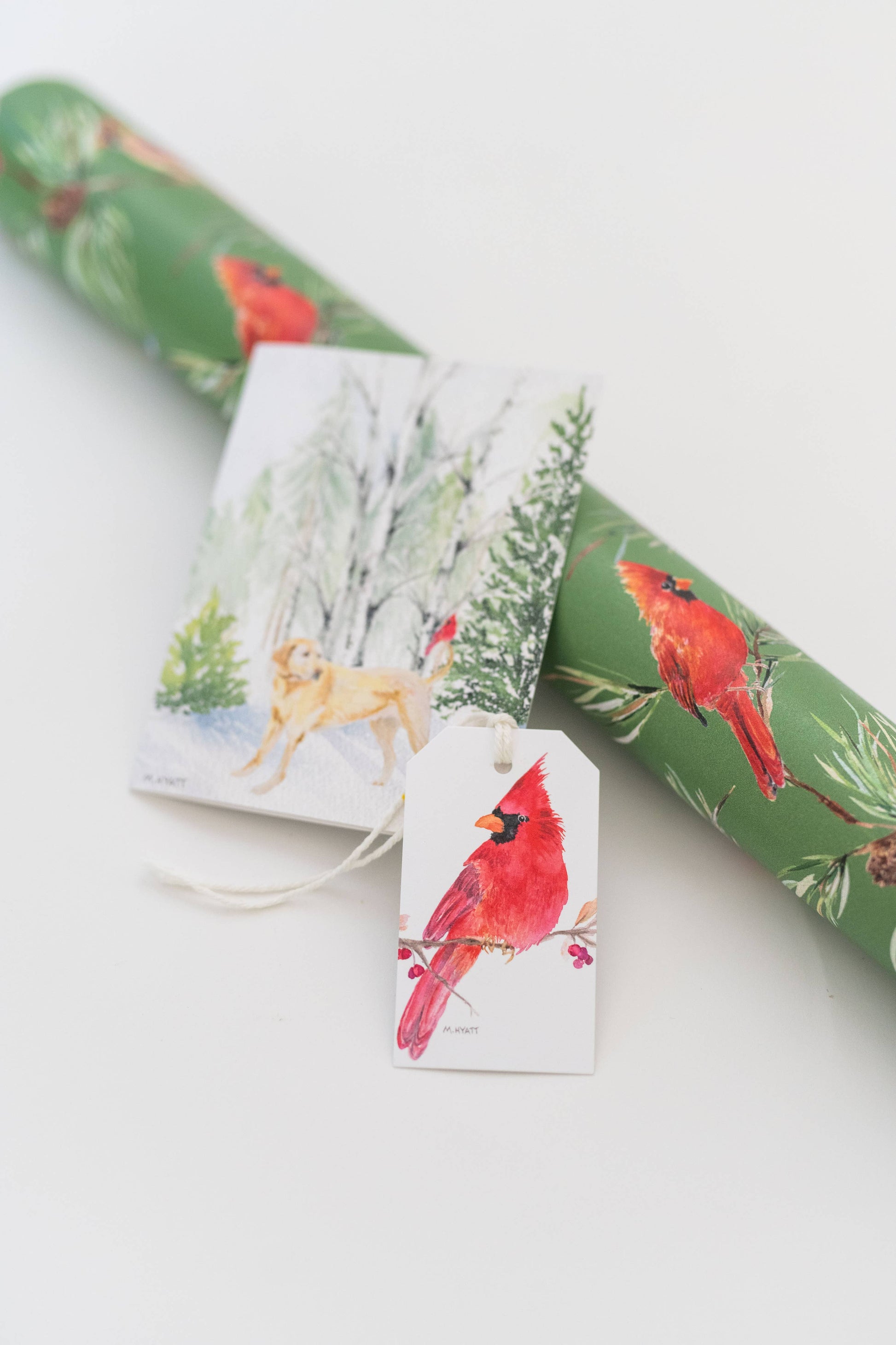 A roll of green holiday wrapping paper with red cardinals and pine branches, alongside a matching gift tag and a small printed card, photographed on a clean white background.