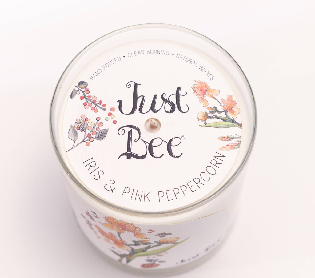 A white scented candle in a clear glass jar with a white lid printed with the brand name 'Just Bee' and the scent 'Iris & Pink Peppercorn,' decorated with floral and berry illustrations.