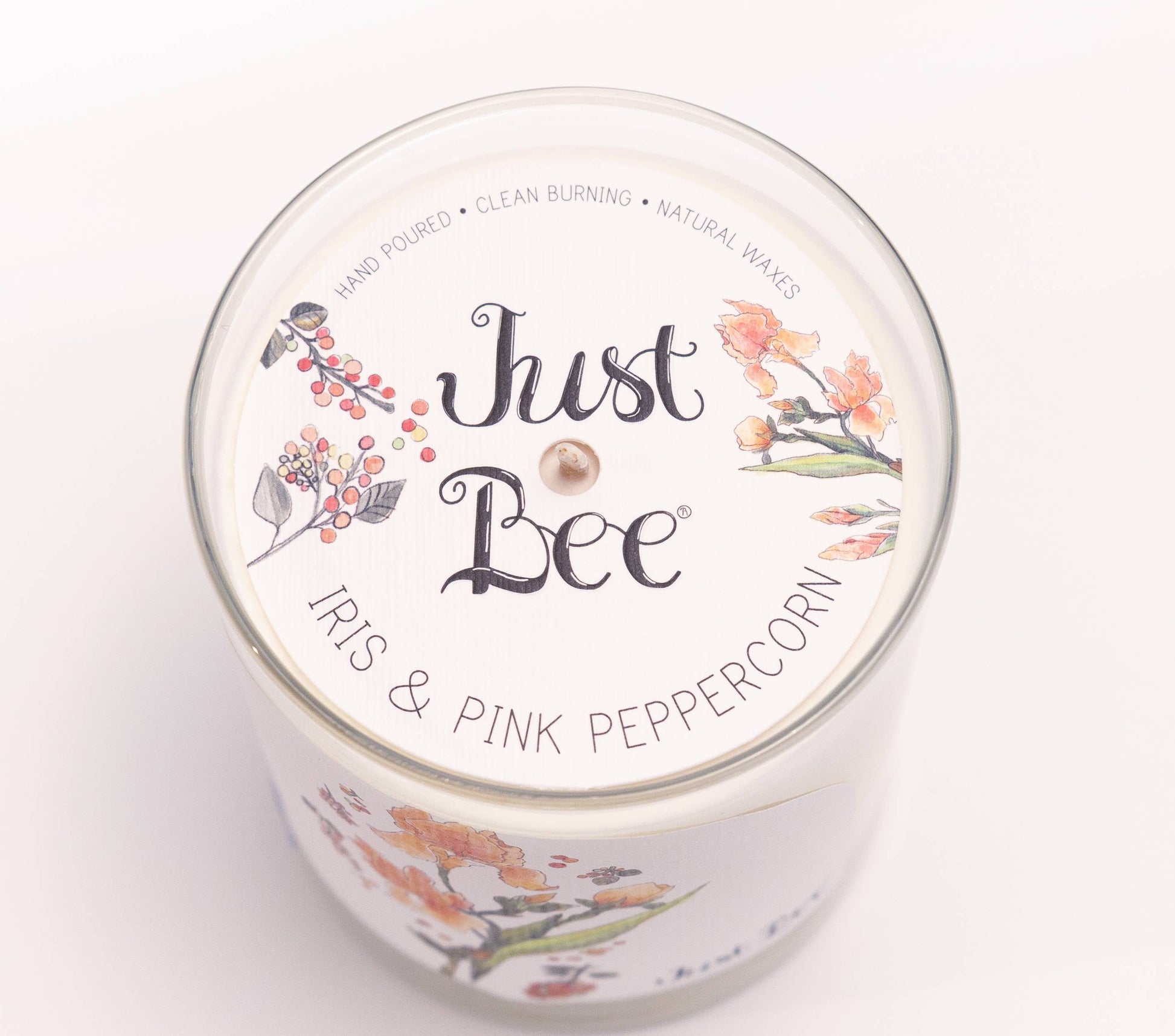 A white scented candle in a clear glass jar with a white lid printed with the brand name 'Just Bee' and the scent 'Iris & Pink Peppercorn,' decorated with floral and berry illustrations.
