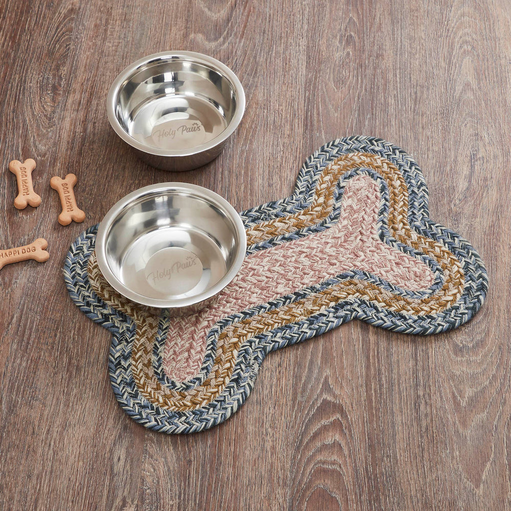 Two stainless steel pet bowls placed on a braided bone-shaped mat with a wood-look floor background. The mat has a braided pattern in gray, tan, and blush pink, and there are small bone-shaped treats nearby.