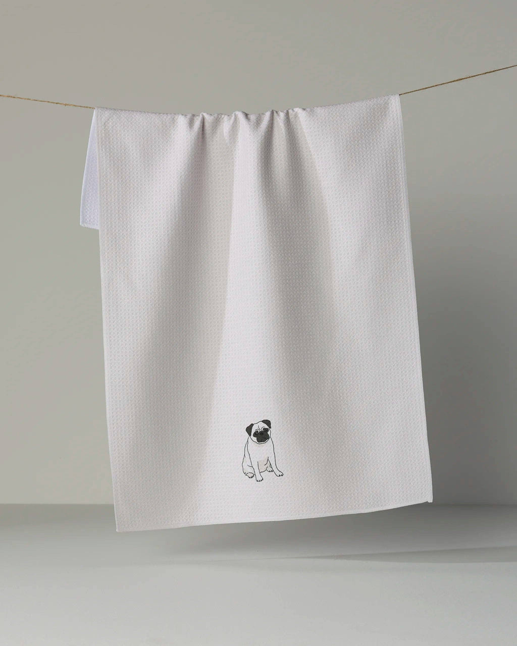 A white waffle-weave kitchen towel with a small black and white pug dog illustration near the bottom, hanging from a thin string against a neutral gray background.