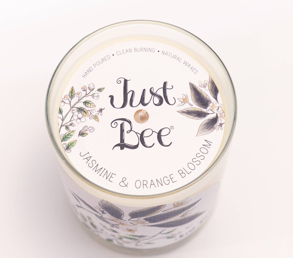 Top-down view of a glass jar candle with a white lid label reading 'Just Bee' and 'Jasmine & Orange Blossom,' decorated with botanical illustrations on a white background.