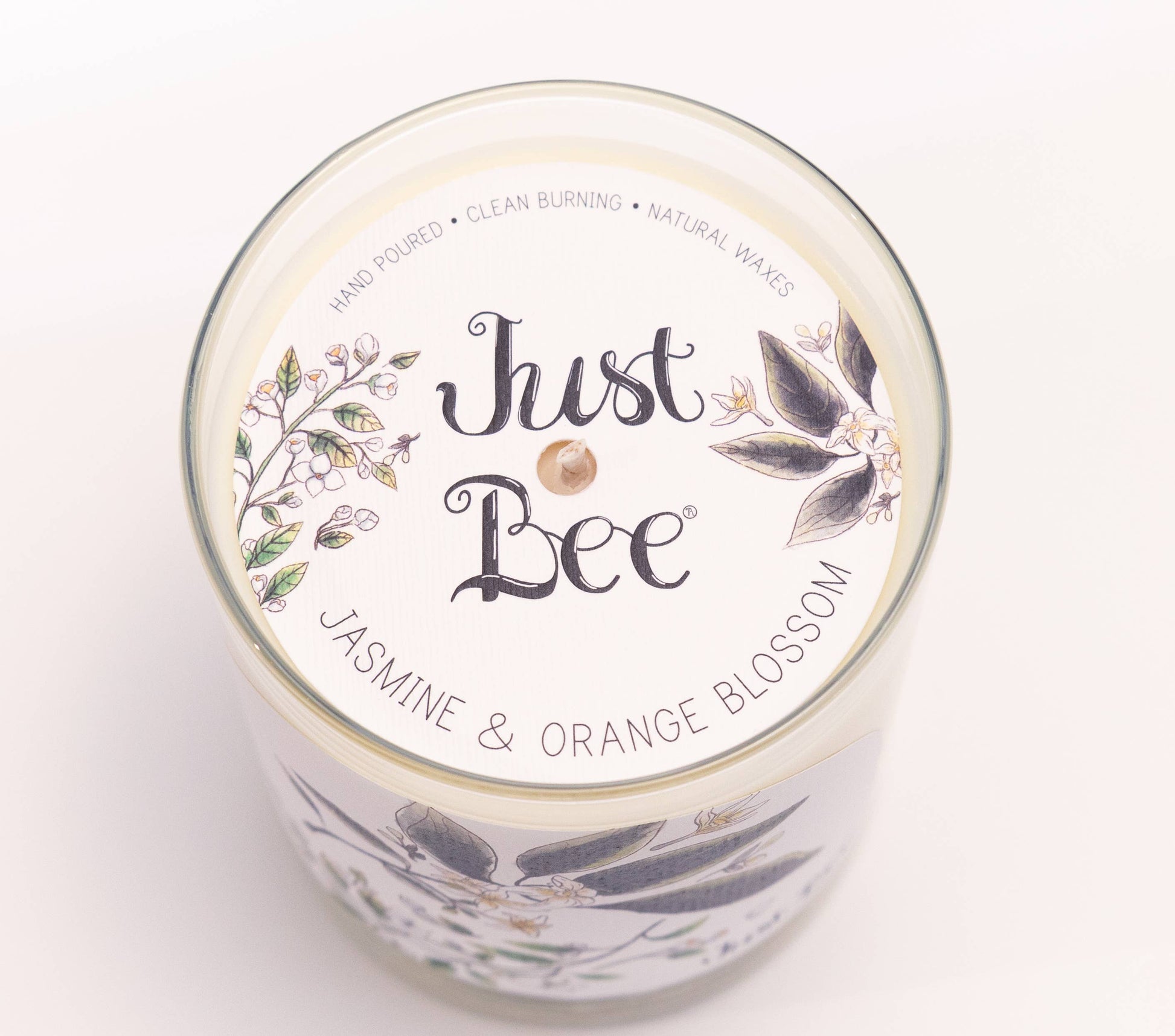 Top-down view of a glass jar candle with a white lid label reading 'Just Bee' and 'Jasmine & Orange Blossom,' decorated with botanical illustrations on a white background.